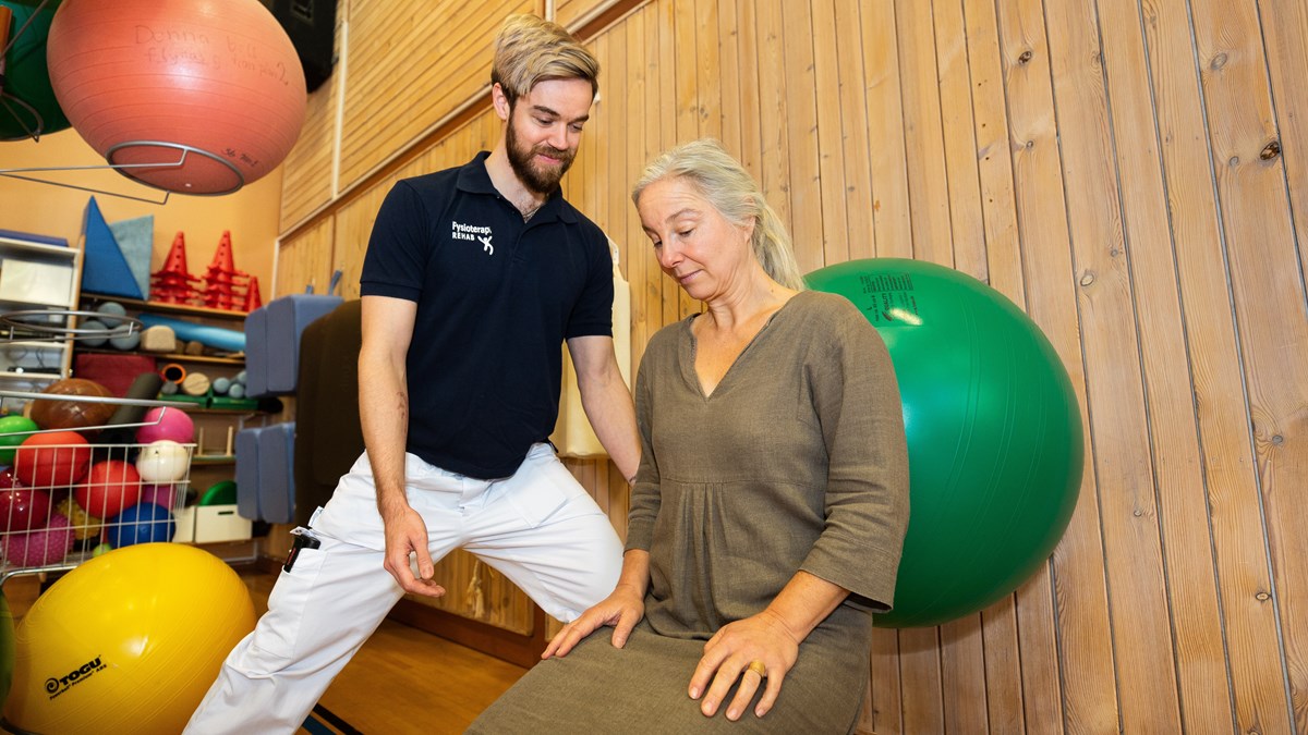En fysioterapeut står i en idrottshall. En patient gör rehabiliteringsövningar med en pilatesboll. 