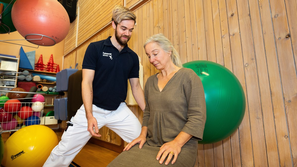 En fysioterapeut står i en idrottshall. En patient gör rehabiliteringsövningar med en pilatesboll. 