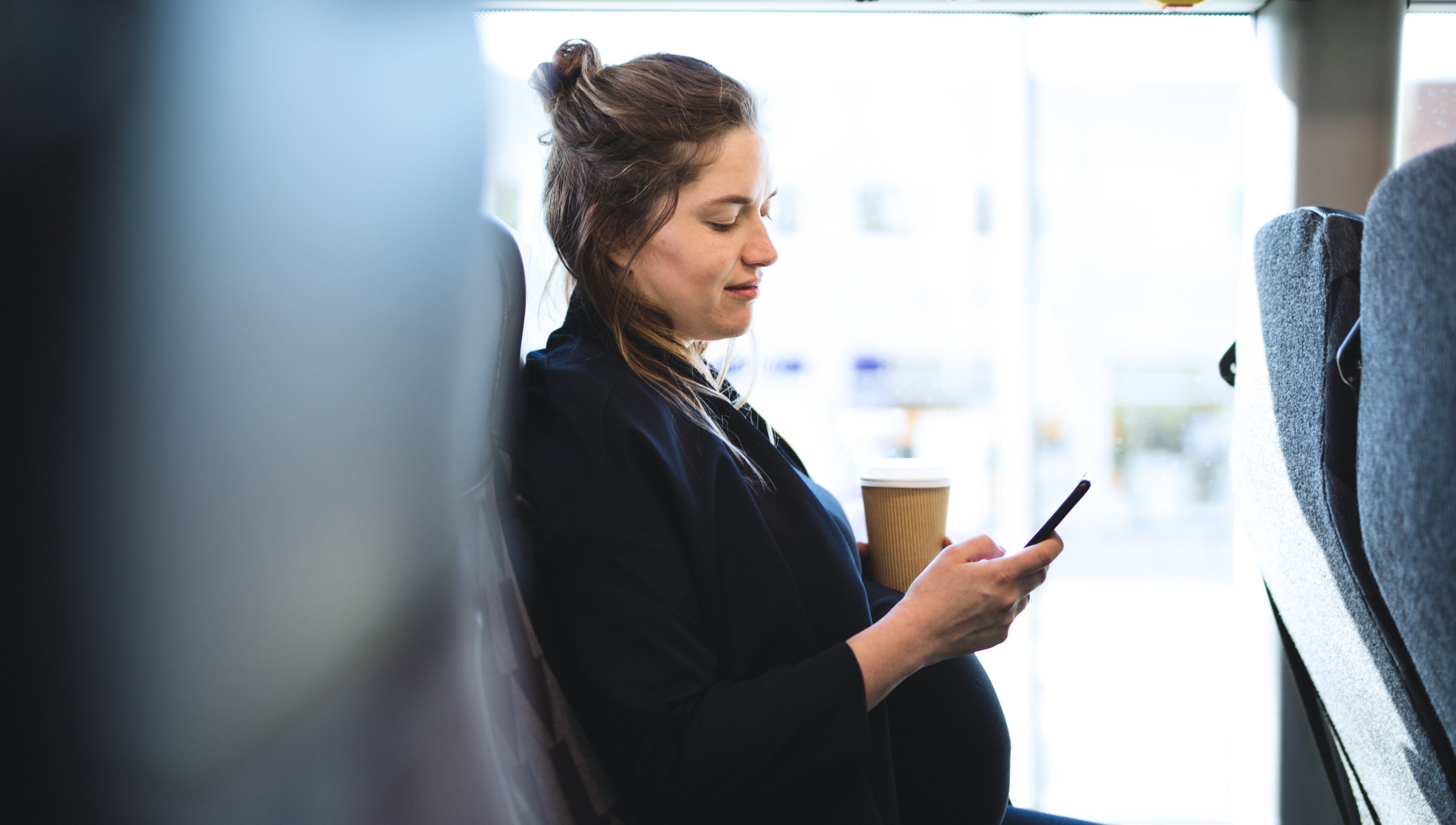 Gravid kvinna på buss tittar i sin mobiltelefon men en kaffe i handen. 
