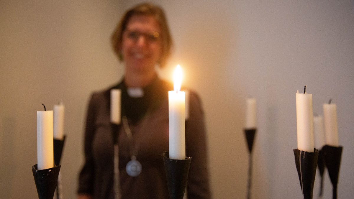 Flera vita ljus, varav ett är tänt. Kvinna i bakgrunden som arbetar inom kyrkan ler.