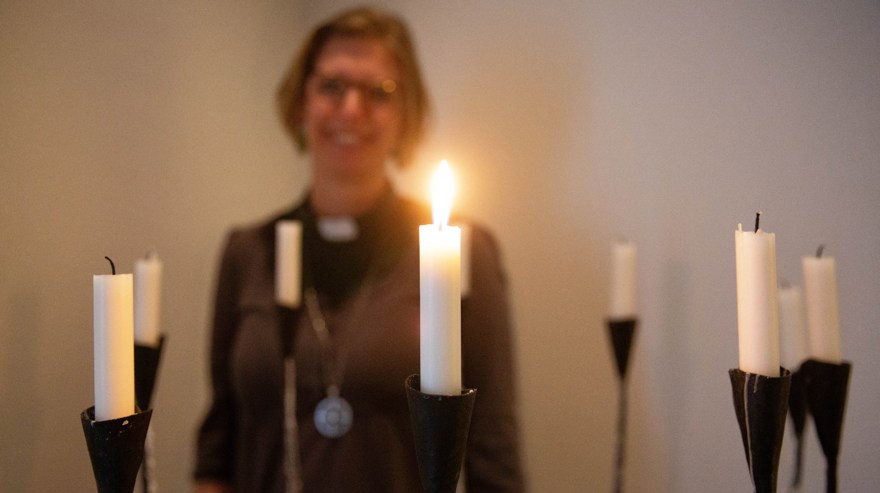 Flera vita ljus, varav ett är tänt. Kvinna i bakgrunden som arbetar inom kyrkan ler.