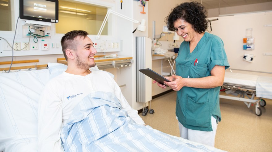 Kvinna i arbetskläder står vid sjukhussäng. I sängen ligger en leende patient.