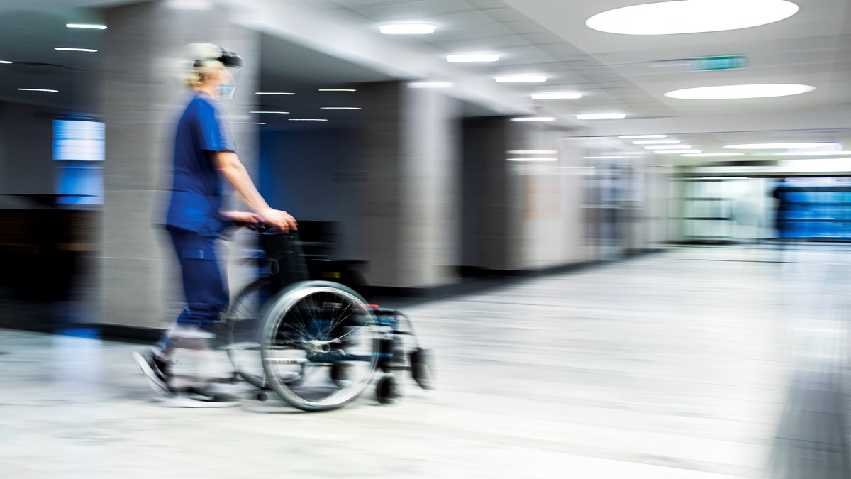 Vårdpersonal i arbetskläder kör rullstol i korridor på sjukhuset.