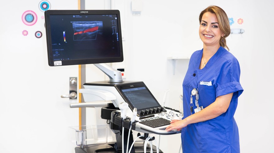 Leende kvinna i arbetskläder står vid en radiologiutrustning med datorskärm.