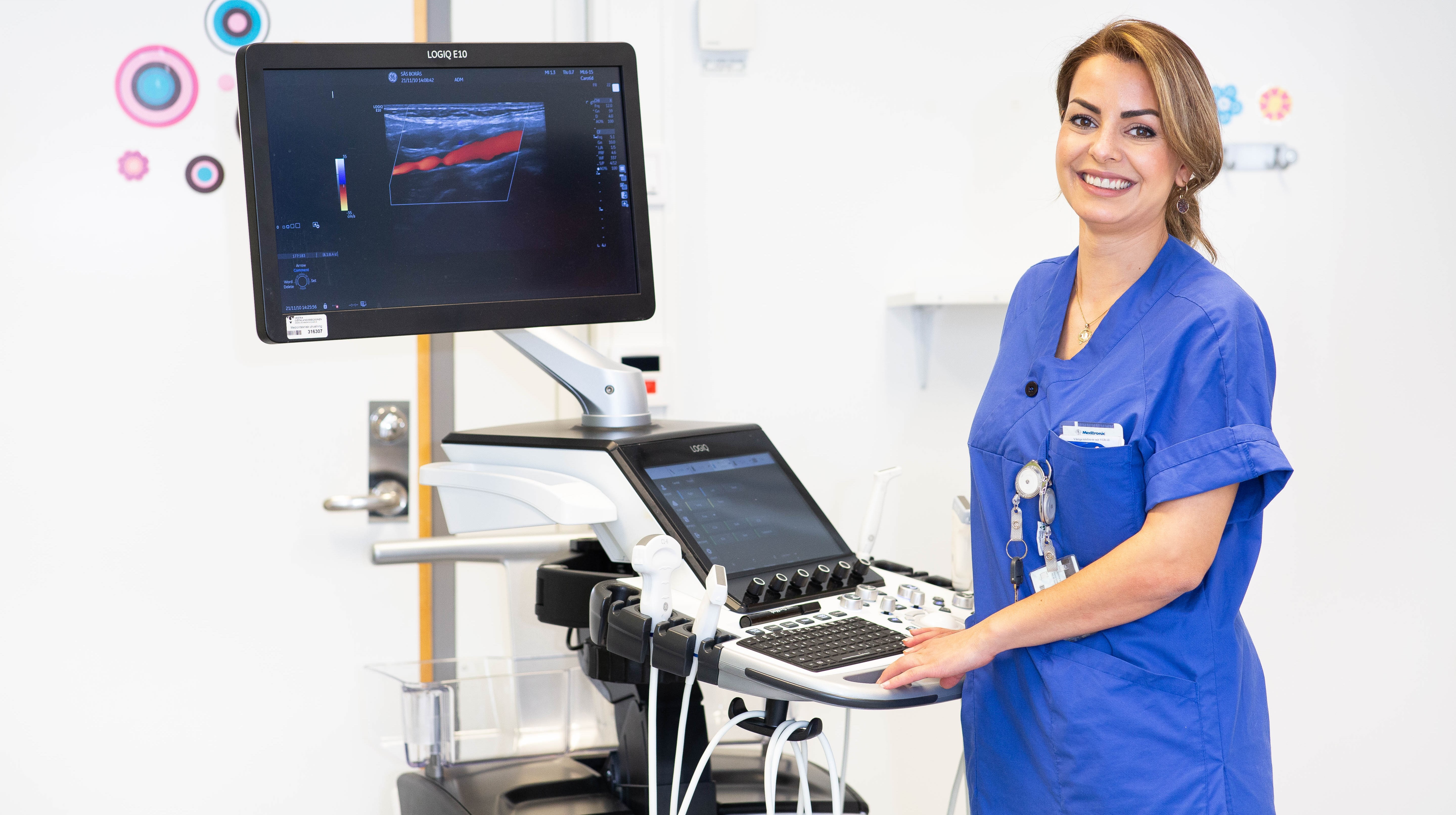 Kvinna i arbetskläder står vid en datorskärm och annan vårdutrustning.